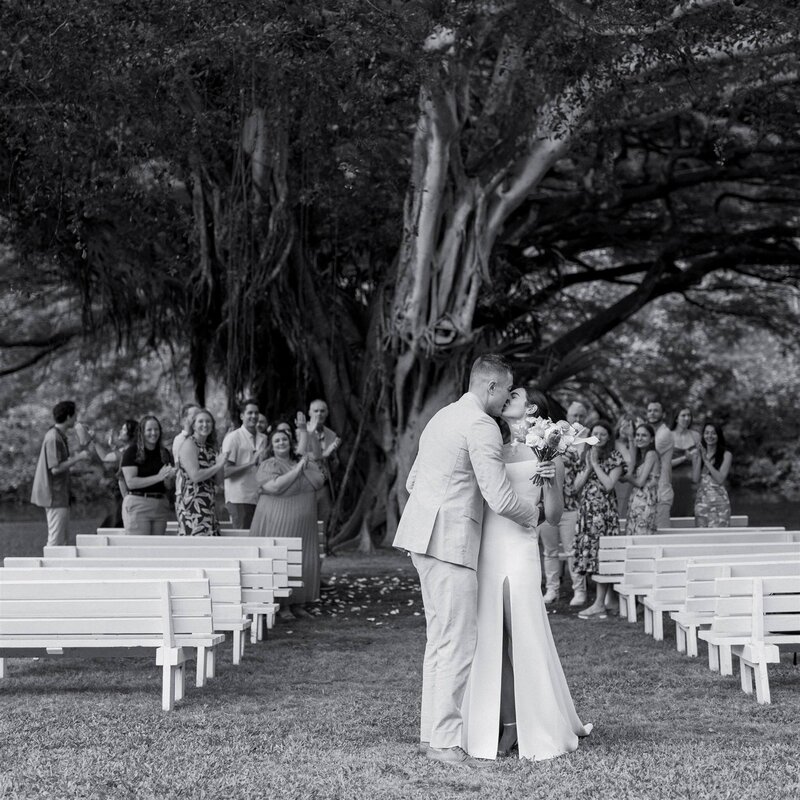 A newlywed couple kissing with wedding guests clapping behind them 