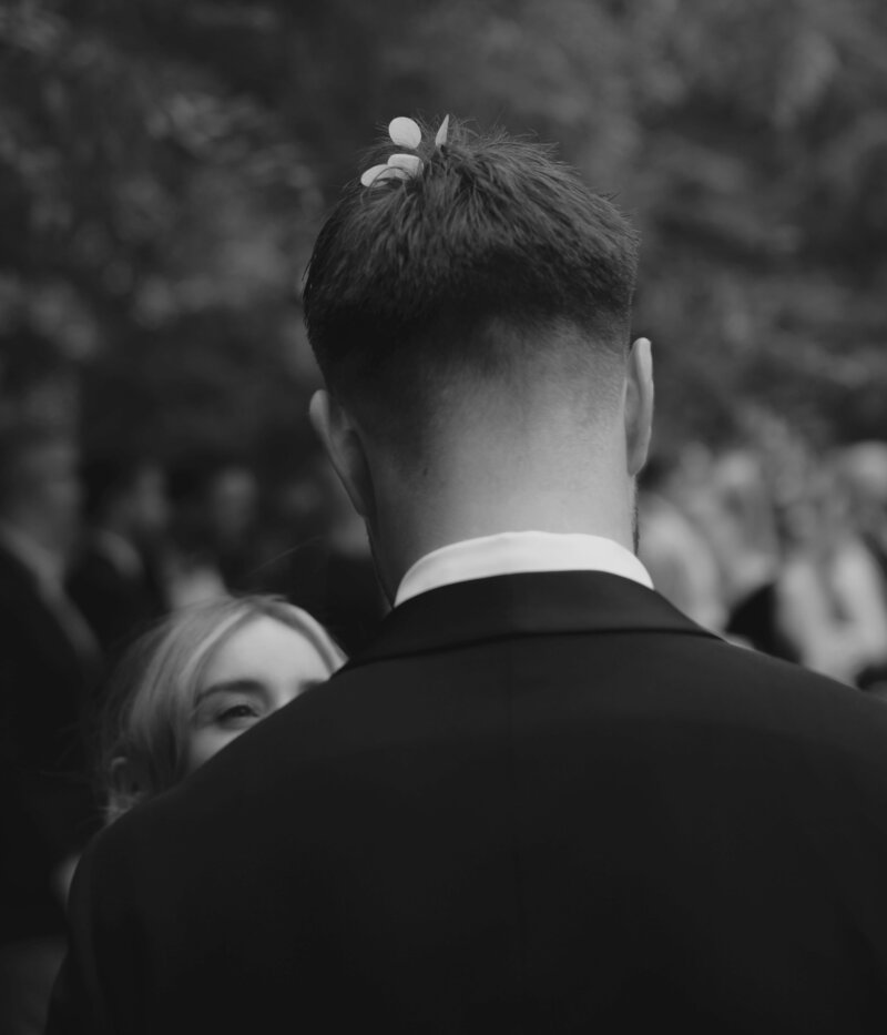 A black-and-white photograph taken at Burnley Gardens in Richmond, capturing a tender moment between the bride and groom before their wedding reception at Cutler and Co. The groom is seen from behind, dressed in a classic black suit, with a few small petals resting atop his neatly trimmed hair, suggesting a recent celebratory moment. The bride is partially visible, her head tilted upward towards the groom, her eyes softly peeking over his shoulder, smiling. The background is blurred, with out-of-focus guests and foliage adding depth to the intimate scene. The monochrome filter emphasizes the emotional connection between the couple, creating a timeless and romantic atmosphere.