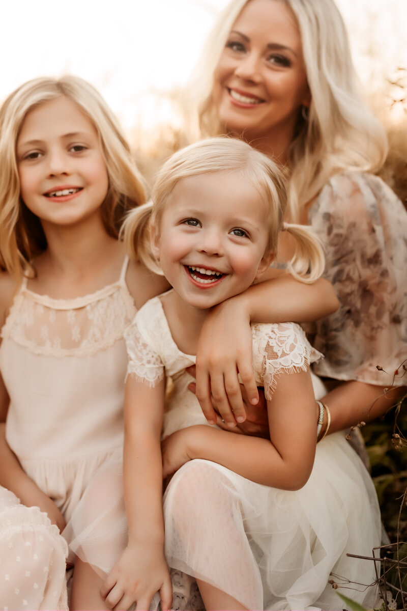 Kansas City Lifestyle Photographer captures little girl smiling big while her older sister has her arm around her and they sit on mom's lap during family photo session.