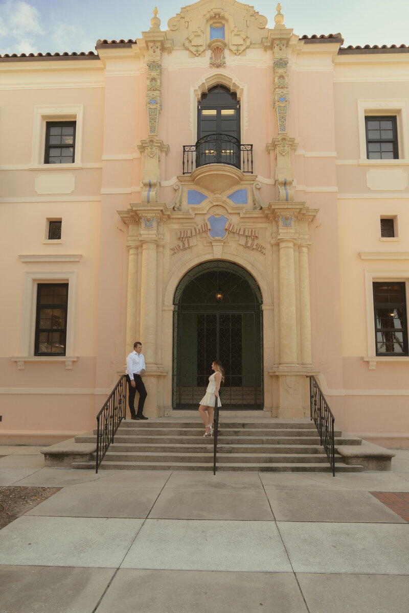 Stylish woman wearing gold geometric sunglasses, white head wrap, and layered gold jewelry, holding a flower in her mouth and Chinese takeout box filled with colorful flowers in a playful editorial-style portrait outdoors.