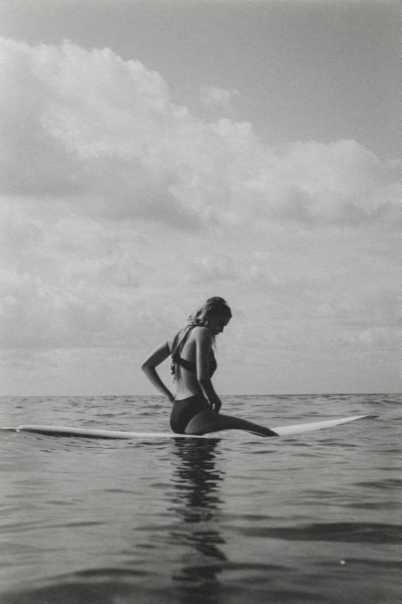 A black and white photo of a woman confidently standing on a surfboard in the ocean, embodying freedom, balance, and empowered entrepreneurship.