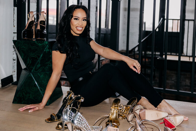 Tiara in a black top and black pants sitting on the ground surrounded by pairs of heels