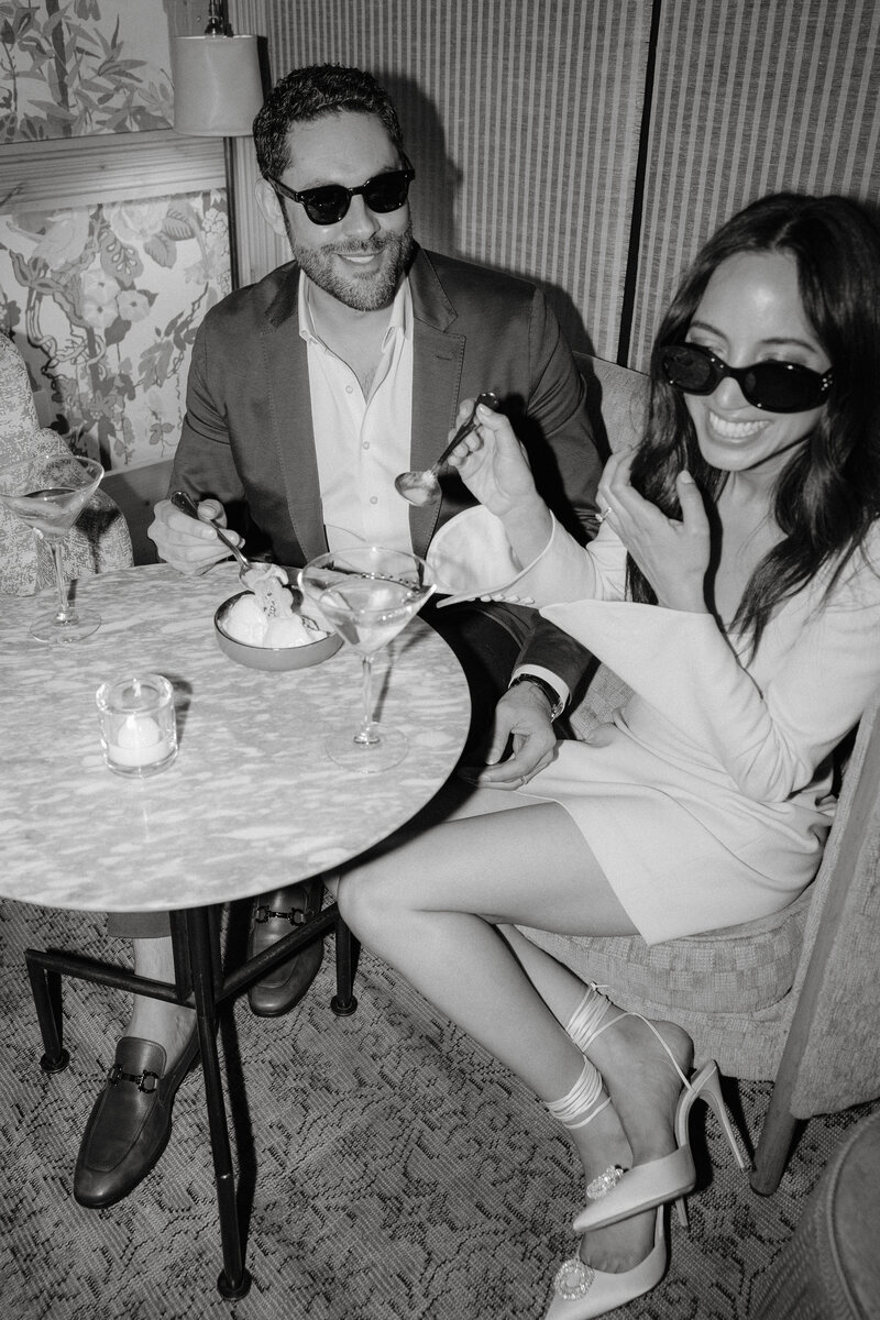 Black and white photo  of man and woman wearing sunglasses sitting at a restaurant table with drinks and ice cream