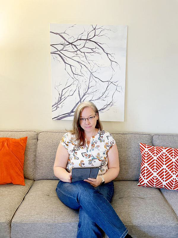 The image shows Sandra sitting comfortably on a gray sofa. She holds a tablet in her hands, which are resting on her lap. Sandra has shoulder-length blonde hair, wears glasses, and is dressed in a white blouse with a butterfly and floral pattern, paired with blue jeans. Behind her, a large canvas with an artwork of a stark tree branch is mounted on the wall. To her sides, bright orange and red patterned throw pillows add a pop of color to the neutral-toned room. Sandra appears relaxed and engaged with her tablet.