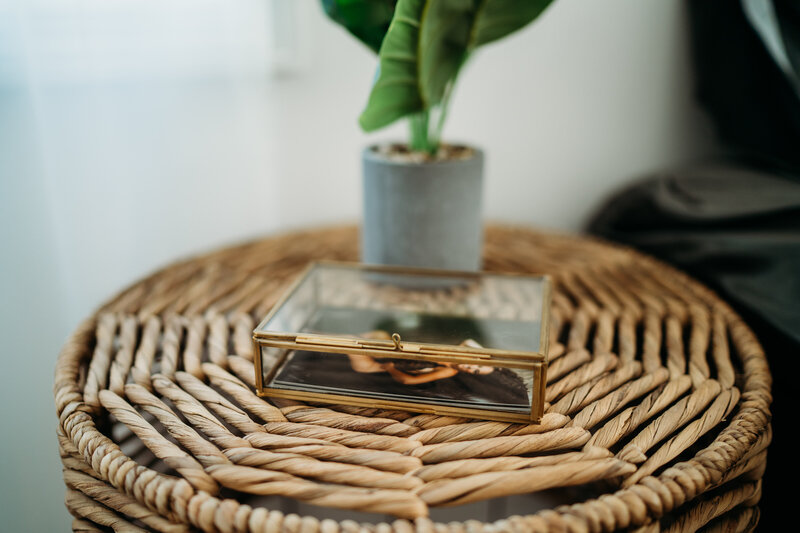 A glass box with luxury boudoir prints sits on a wicker bedside table with a house plant