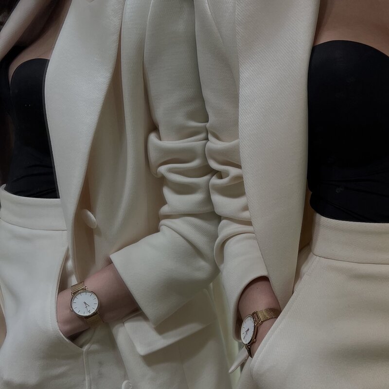 Two people in elegant, cream-colored suits with black tops. Each wears a gold watch. The scene exudes sophistication and style.