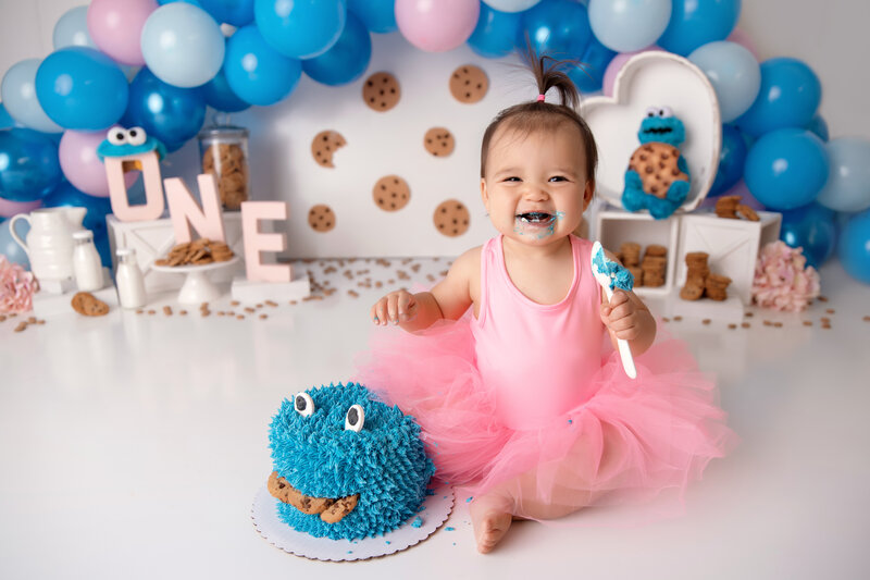 one-year old girl eating a pink donut and sitting in front of her birthday cake as part of her cake smash session