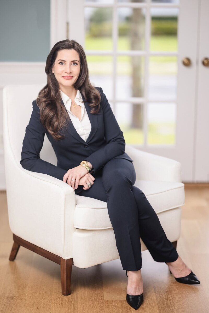 Corporate branding portrait of a woman in a navy suit sitting in a white chair with natural light background, Long Island business headshots.