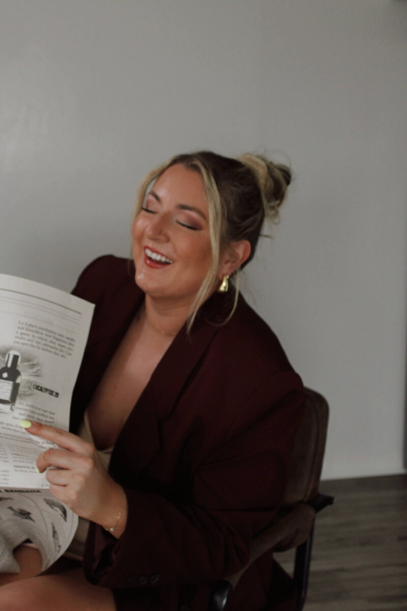 Mikayla Leier, the founder of The Aesthetic Edit, a company specializing in aesthetic website design and marketing for female owned small businesses, shares a joyful moment reading a newspaper. Her approachable demeanor reflects the personalized service offered.