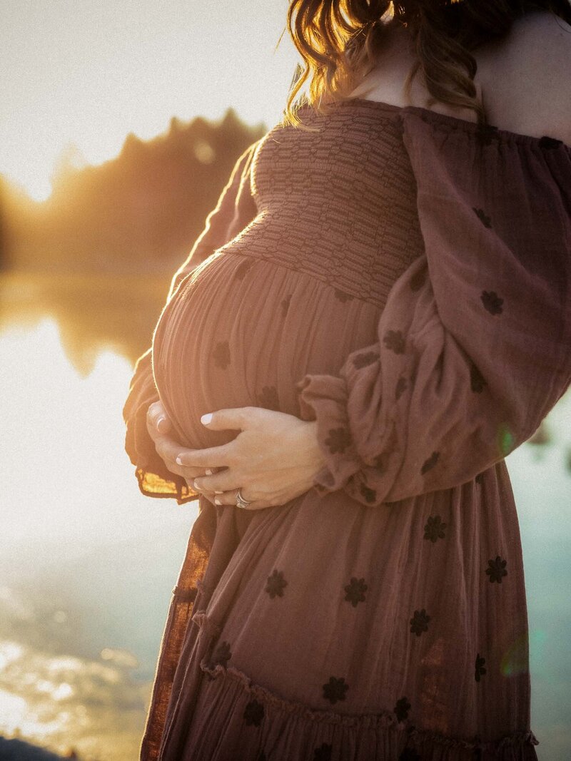 Close-up of a pregnant mother in a rust-colored dress holding her baby bump in warm sunset light.