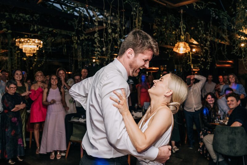 a groom and bride laugh while doing first dance in front of vines at 5th street restaurant during their wedding reception in christchurch