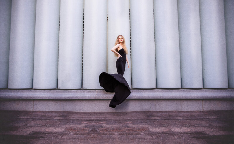 High school senior in a black designer gown flipping her skirt during a stylized portrait session in front of modern architectural columns