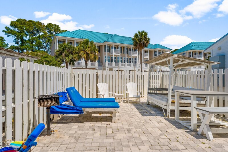 Sunny fenced-in backyard patio at Tropical House with blue loungers, Adirondack chairs, and a grill, just minutes from Panama City Beach.