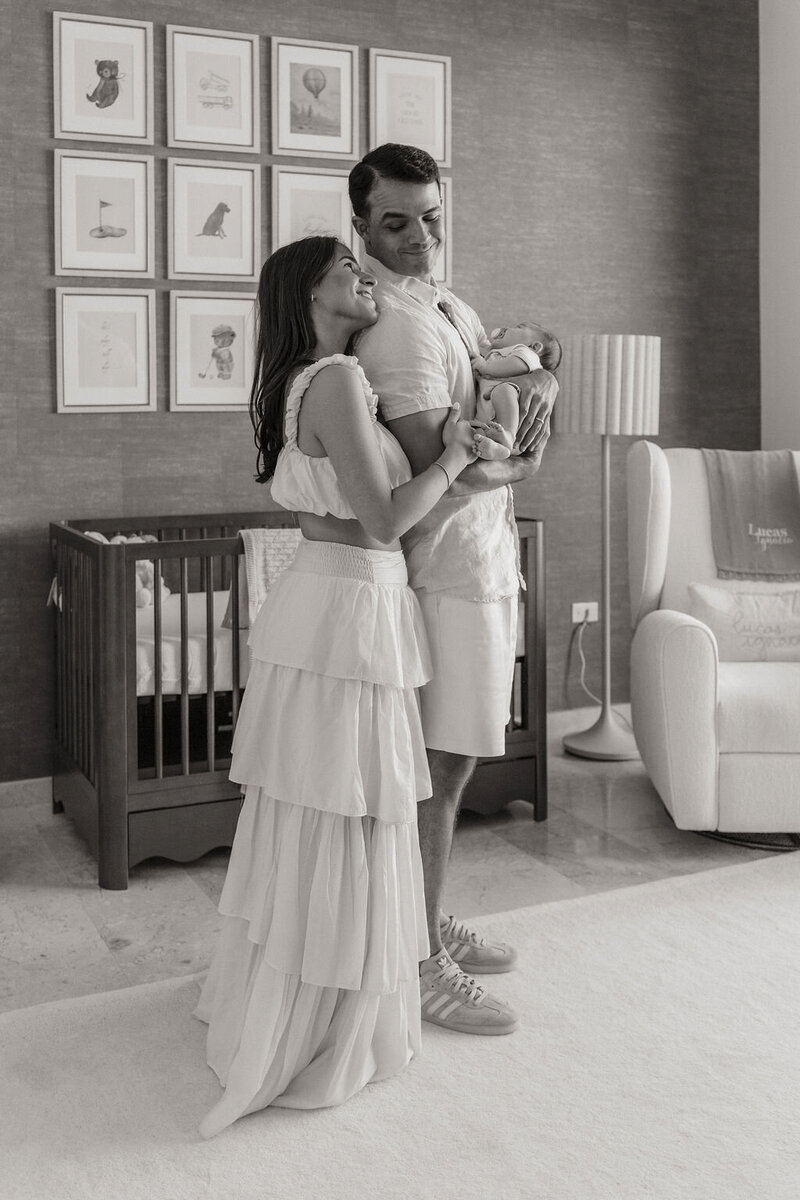 Black and white photo of mom, dad, and baby in nursery with framed animal prints above crib.