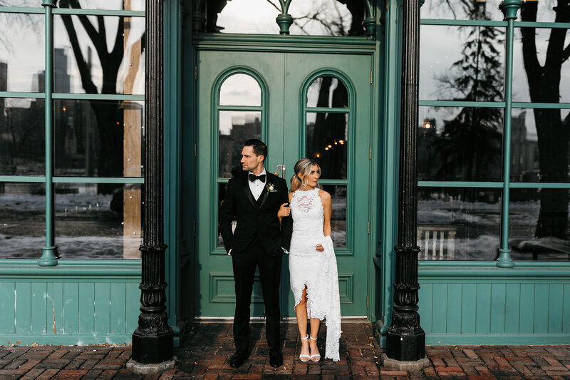 Bride and Groom standing in front of door with arms linked looking the opposite direction without smiling