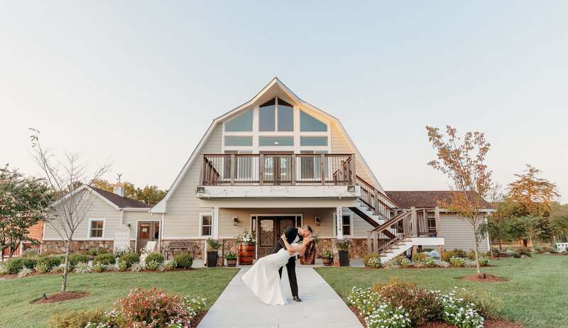 A couple shares a romantic dance pose in front of an elegant outdoor wedding venue in Virginia, with a central staircase leading to a balcony. The surrounding garden is well-manicured with flowers and trees under a clear sky, making for perfect barn wedding pictures.