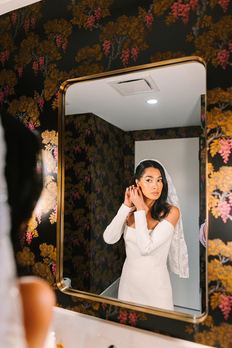 Bride adjusting earrings in the mirror with colorful wallpaper on the wall around her
