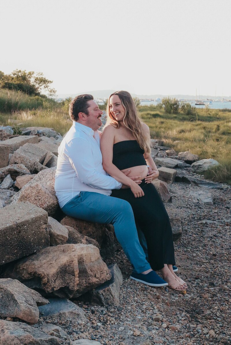 Couple sitting on rocks on beach smiling at each other during maternity photoshoot.
