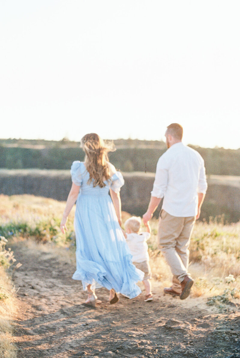 Husband wearing white dress shirt and khaki pants,  wife wearing blue flowy long dress and blonde curled hair, and two year old son wearing white dress shirt and khaki shorts walking away from view walking down a dirt pathway lined with wildflowers on mountainside ledge by Portland Newborn Photographer Emilie Phillipson Photography.