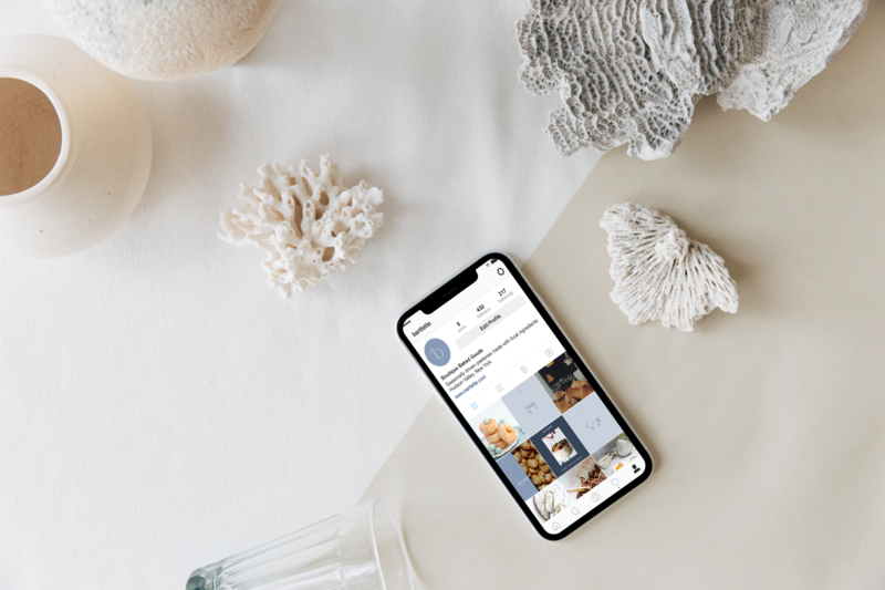 iPhone with a social media profile of a bakery on a table with pieces of coral and a glass