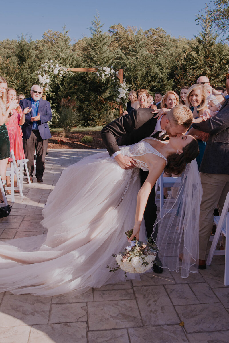 Amidst the enchanting backdrop of an outdoor wedding venue in Virginia, a bride and groom share a dip kiss. The bride in her flowing white gown with bouquet contrasts beautifully with the groom's dark suit, as guests clap beneath a floral arch surrounded by lush greenery.