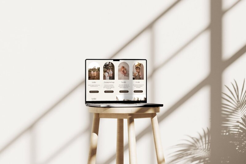 Open laptop on a simple wooden stool against a white wall with dramatic window shadows, displaying a photographer’s services page with four arched images for family, maternity, newborn, and lifestyle sessions.