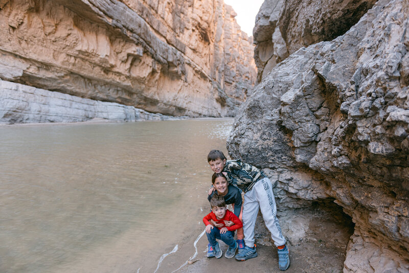 Editorial-style family photo of three children smiling against canyon cliffs and river water in Big Bend National Park, captured in a fine art, documentary style.