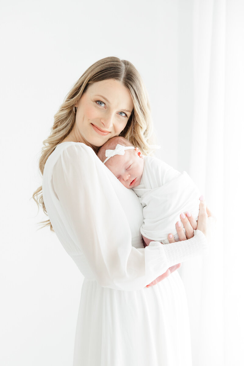 Mother in flowing white dress holds her newborn baby girl, swaddled and wearing a bow, during newborn photography session