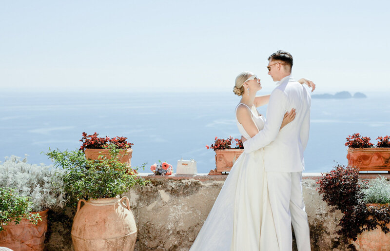 A stunning wedding couple dressed in all white, basking in the sunshine of Positano, with the picturesque coastal town and vibrant blue sea as their backdrop.