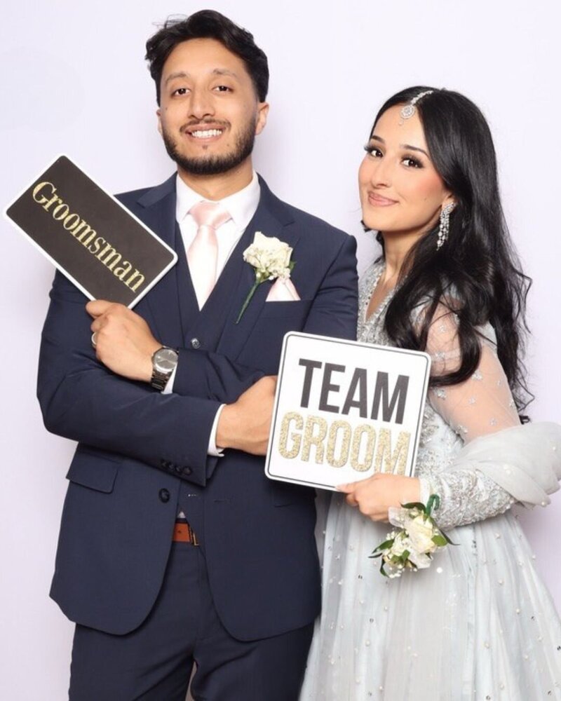 Wedding guests posing with “Groomsman” and “Team Groom” signs in coordinated pastel outfits at photo booth.