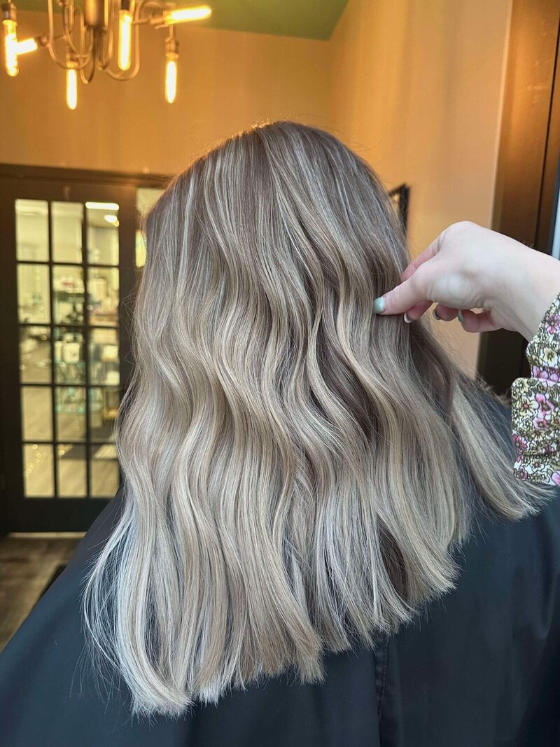 A back view of a woman with soft, wavy hair, with a stylist's hand gently adjusting it, showcasing a natural and elegant hairstyle created by 212 Salon, Spa, & Barbershop.