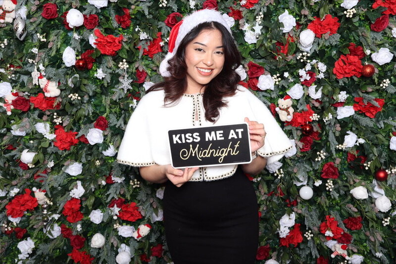 Woman wearing a Santa hat holding a “Kiss Me at Midnight” sign in front of a red and white Christmas floral backdrop at AR Photobooth.