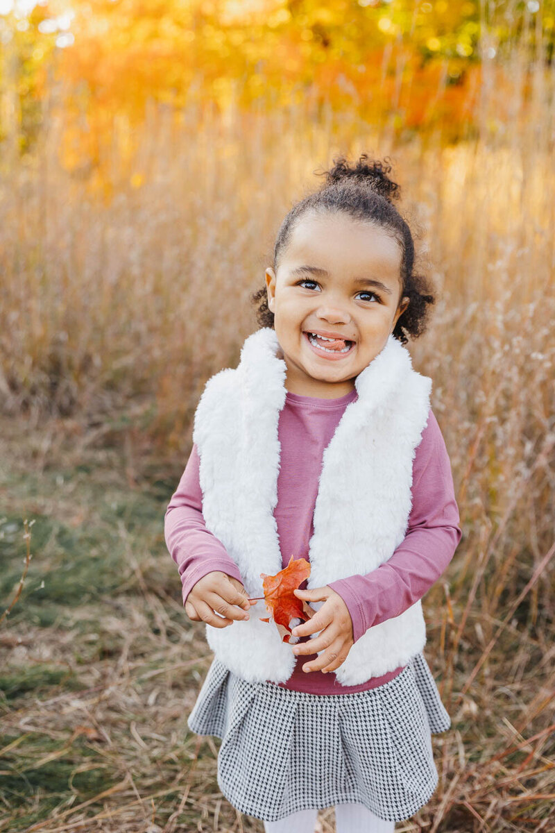A child smiles, clutching a red leaf in a grassy field filled with autumn's orange hues. Captured during a fall portrait session in High Park