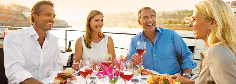 Four adults sitting at an outdoor waterfront table, enjoying a meal and glasses of rosé wine while smiling and talking. Sunlight reflects off the water in the background, and the table is set with flowers, food, and drinks.
