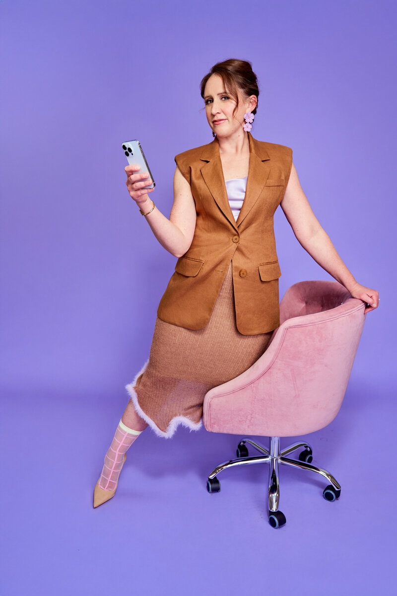 Jaime Bartol, leadership coach, sitting confidently with smartphone in hand against a purple background and pink chair