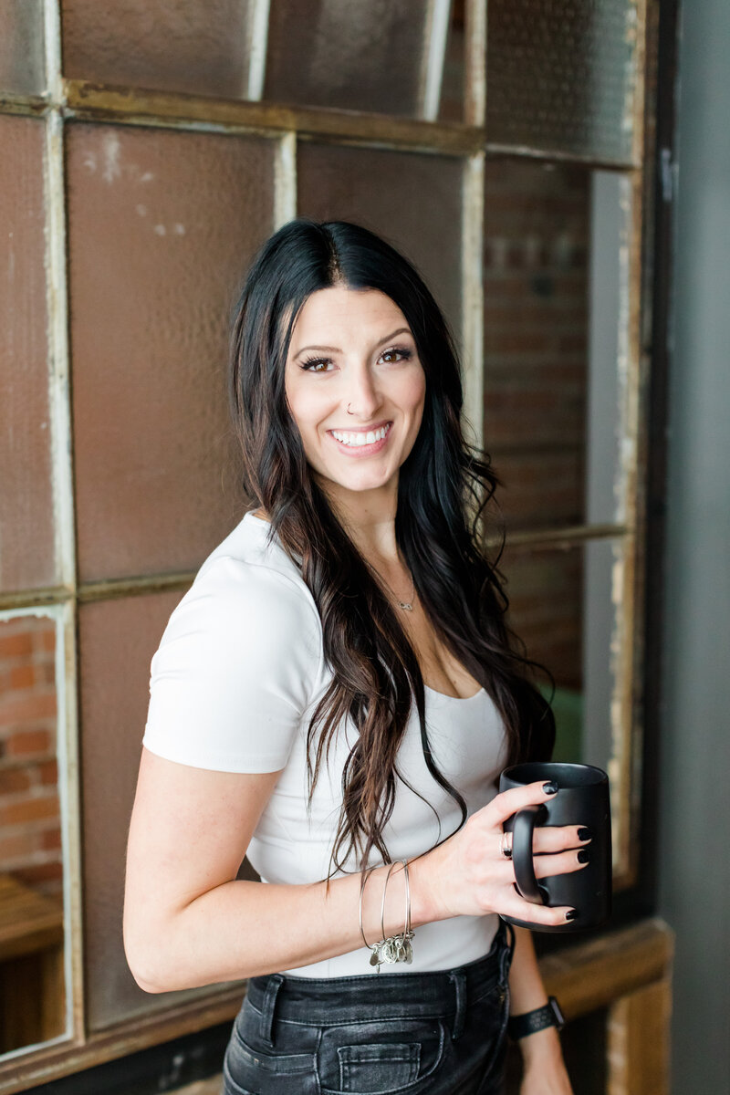 Allie Clark, founder of Allie Clark Designs, smiling and holding a black coffee mug in a modern studio setting — featured on the ACD contact page.