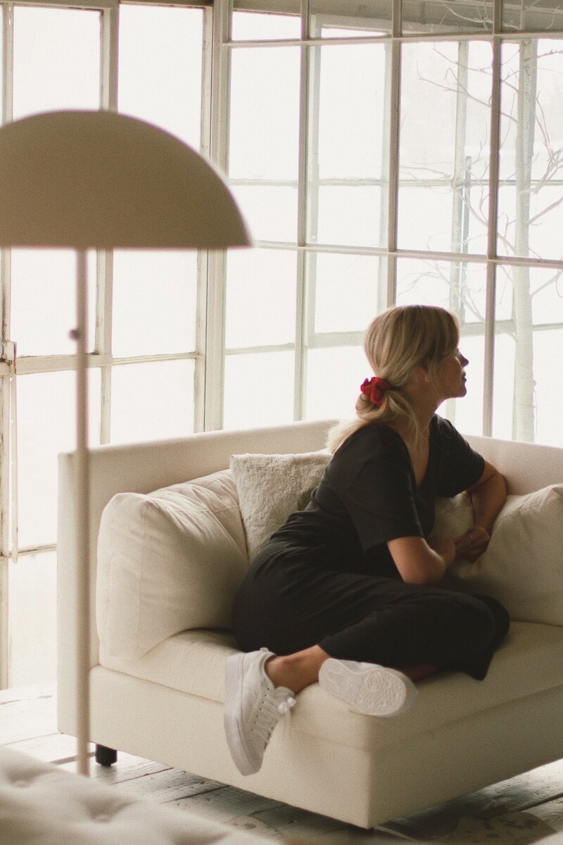 A woman in a black outfit sits contemplatively on a white sofa, with natural light streaming through large windows. The room feels peaceful and airy.