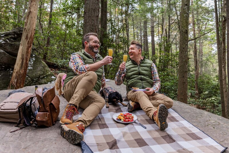 Two grooms sitting on a picnic blanket are smiling at each other cheering with mimosa's after sharing their vows under a waterfall.  They are wearing matching outfits with a plate of fruit between them and a dog laying nearby.