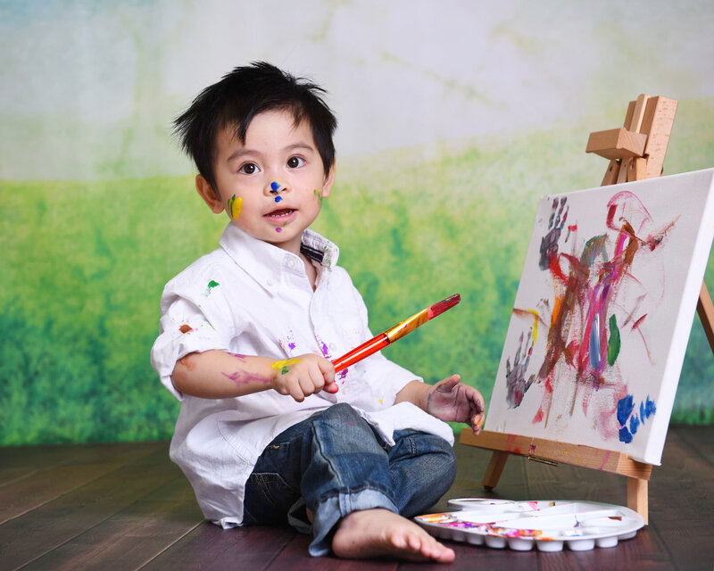 A toddler boy with paint on his face and shirt, holding a brush during an outdoor-themed painting photo shoot. The background features a green grassy vibe with a wooden floor.