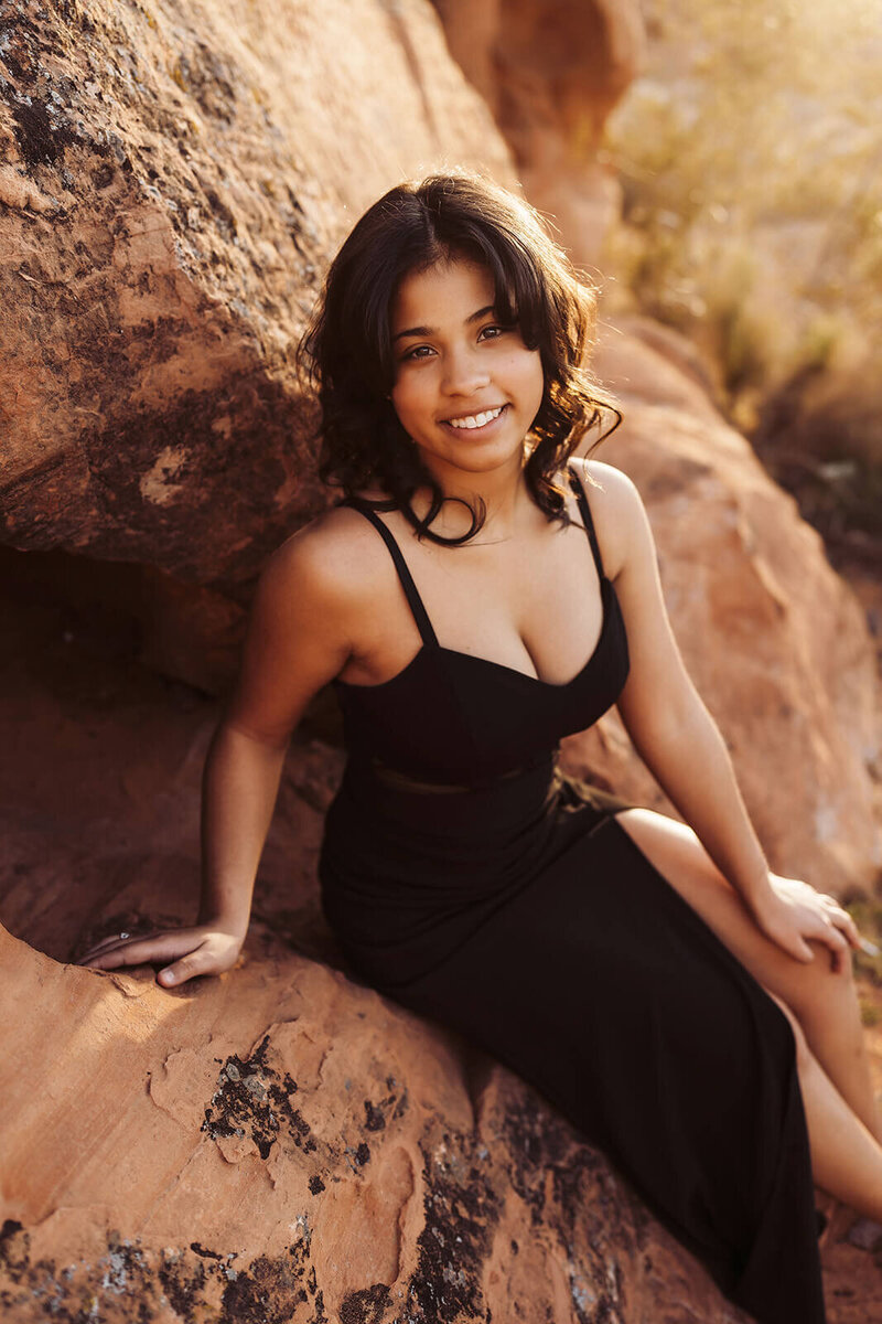 The girl in a black dress is sitting on a rock in the middle of the desert and smiling