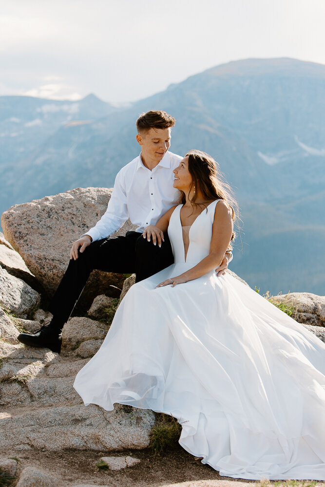 Bride and groom sit on a rock and smile at eachother in Rocky Mountain National Park, Estes Park, Colorado, capturing a joyful and carefree moment during their wedding.