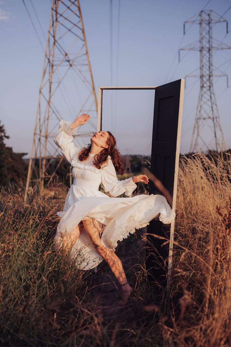 Young, red-headed woman poses in the wind wearing a flowy white dress in a field with a white door and powerlines.