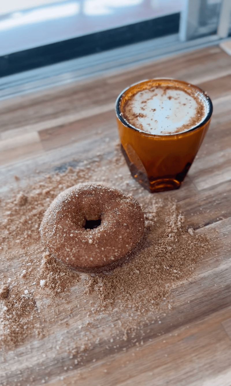 A cinnamon donut and a frothy glass of coffee sit invitingly on the wooden table, surrounded by scattered cinnamon sugar, capturing the cozy essence of a Temple, Texas coffee shop.