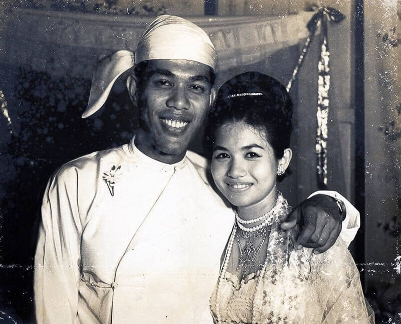 Vintage black and white photo of Nita Tin and her husband Pe on their wedding day in Burma, elegantly dressed in traditional Burmese clothing under a tent, both smiling.