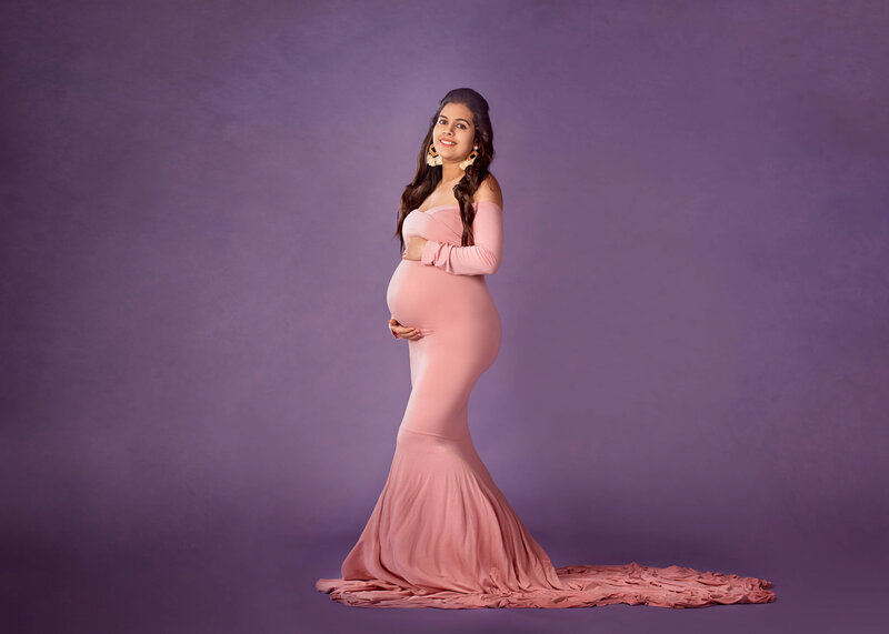Radiant maternity portrait of an expectant mother in a flowing pink gown against a soft purple studio backdrop, captured in Overland Park, Kansas. A timeless choice for families seeking elegant and personalized maternity photography in the Kansas City metro area.