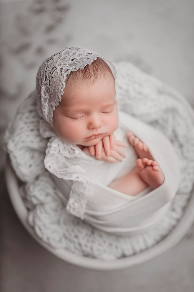 Newborn baby swaddled in white fabric wearing lace bonnet laying on white yarn blanket in a white bowl