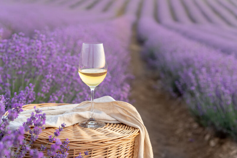 A glass of white wine sits on a woven basket draped with a cloth in a lush lavender field. The purple blooms stretch into the distance under a soft light, creating a serene and picturesque scene.