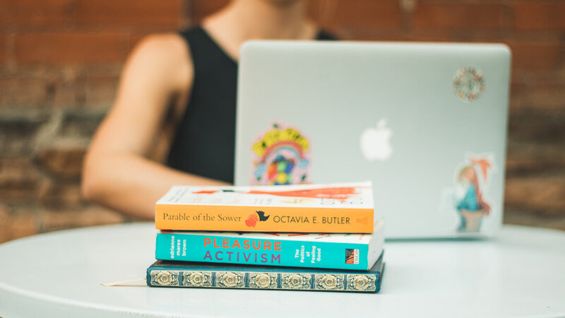 In the front of the image, there's a stack of books: Parable of the Sower by Octavia E. Butler, Pleasure Activism by Adrienne Maree Brown, and a blue and gold journal. Blurred in the background, Kaitlyn Hickmann — founder of Unstoppable Copy — is  sitting with her laptop open.