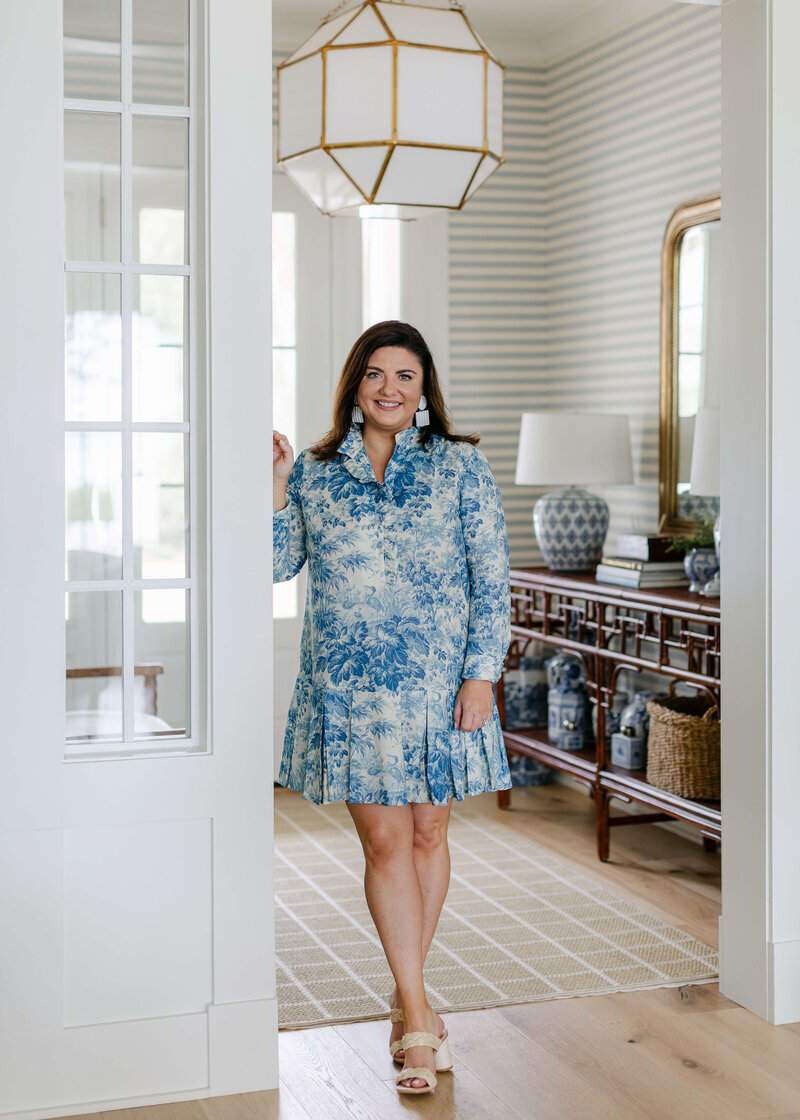 Tiffany is standing in a blue floral dress leaning against a white wall.