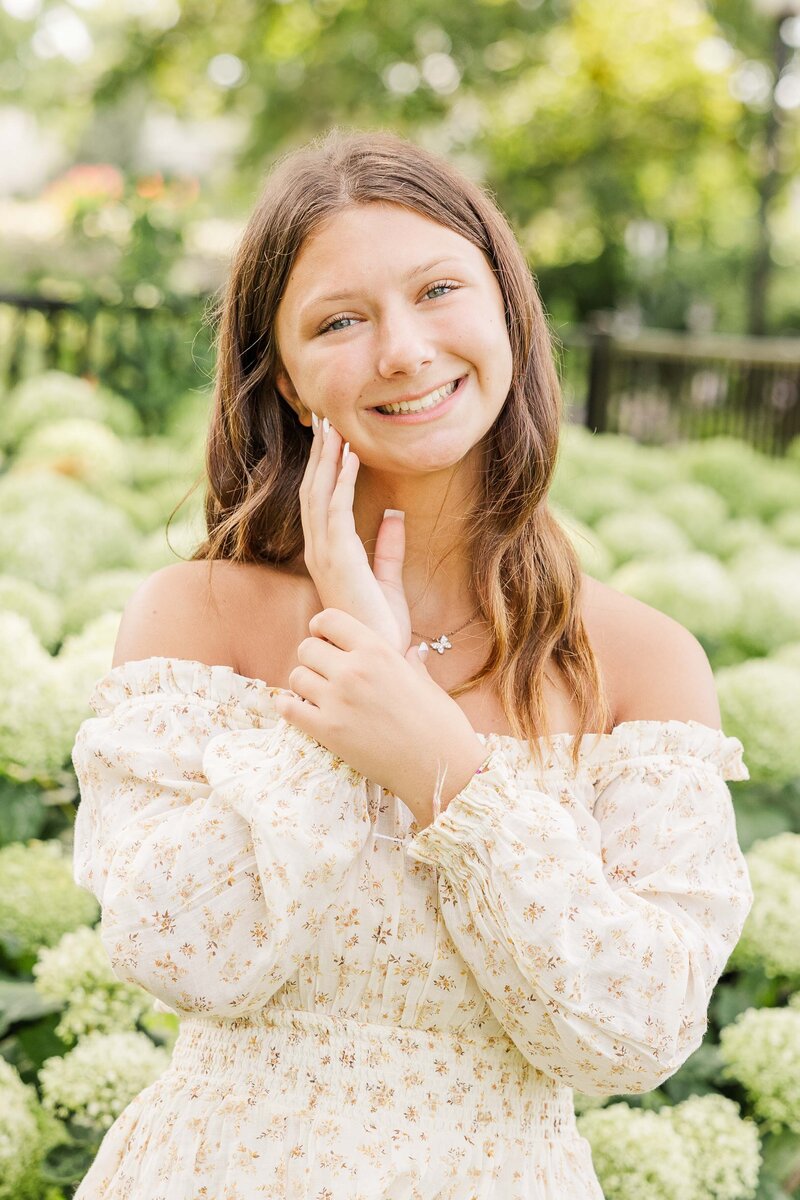 Waukesha Senior Photographer at Frame Park in Waukesha Wisconsin.  Senior holding her face wearing floral dress in front of hydrangea bush in Frame Park, Waukesha.